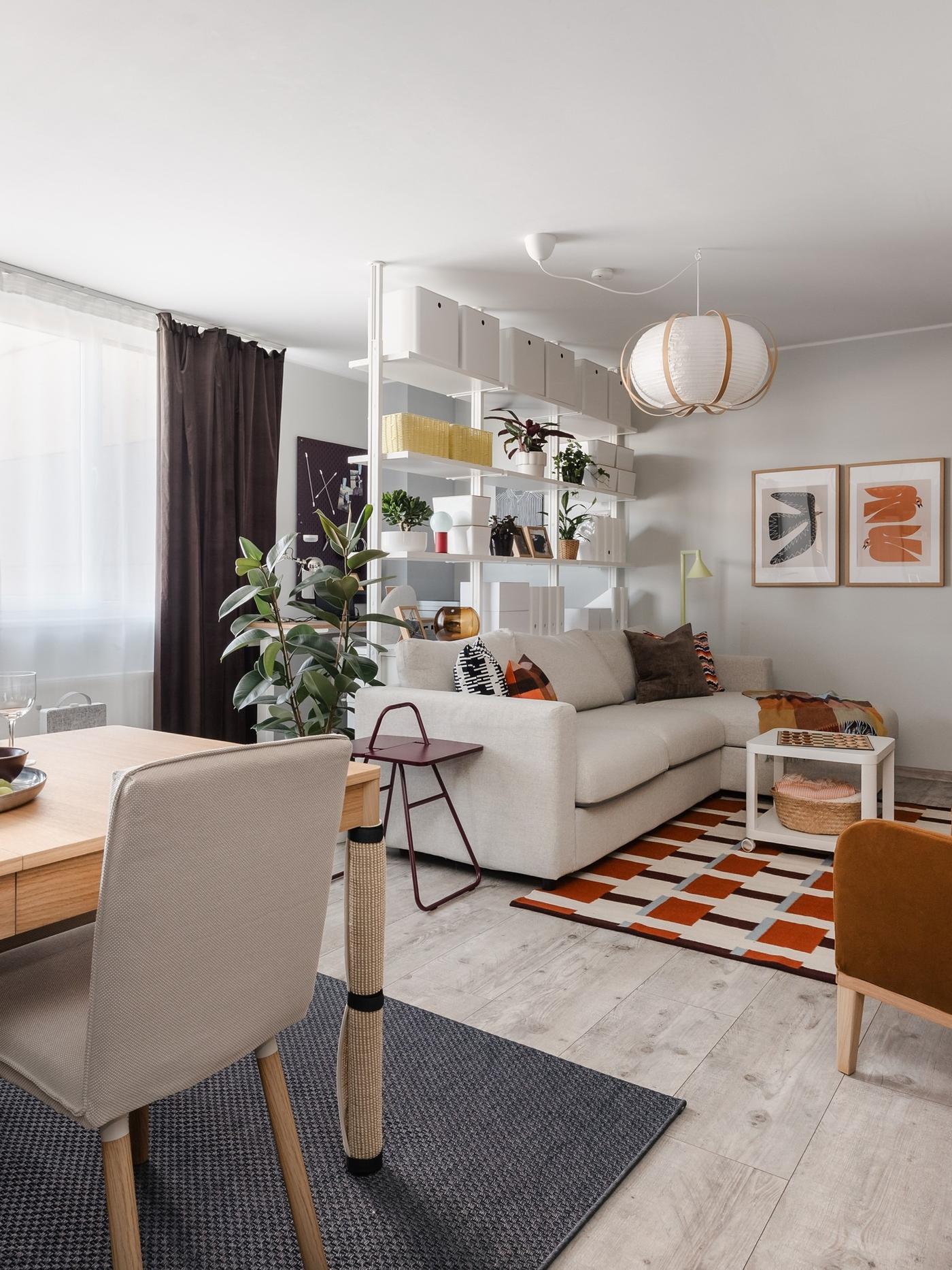 Bright living room after the makeover, with a beige sofa, open shelving, layered rugs, plants and a dining area.