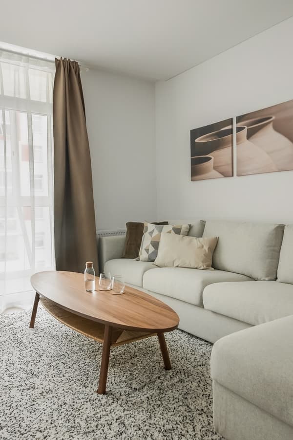 Bright living room featuring a light beige sectional sofa with geometric and neutral-toned pillows. A wooden oval coffee table with glasses and a bottle sits atop a soft, speckled rug. Natural light streams through sheer white curtains beside a brown drape. Above the sofa, a minimalist diptych artwork of ceramic vases adds visual interest.