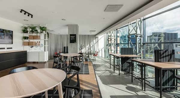 Bright modern coworking space at IKEA with round and high tables, black and red chairs, large floor-to-ceiling windows, indoor plants, and minimalist shelving, combining natural light with clean Scandinavian design.