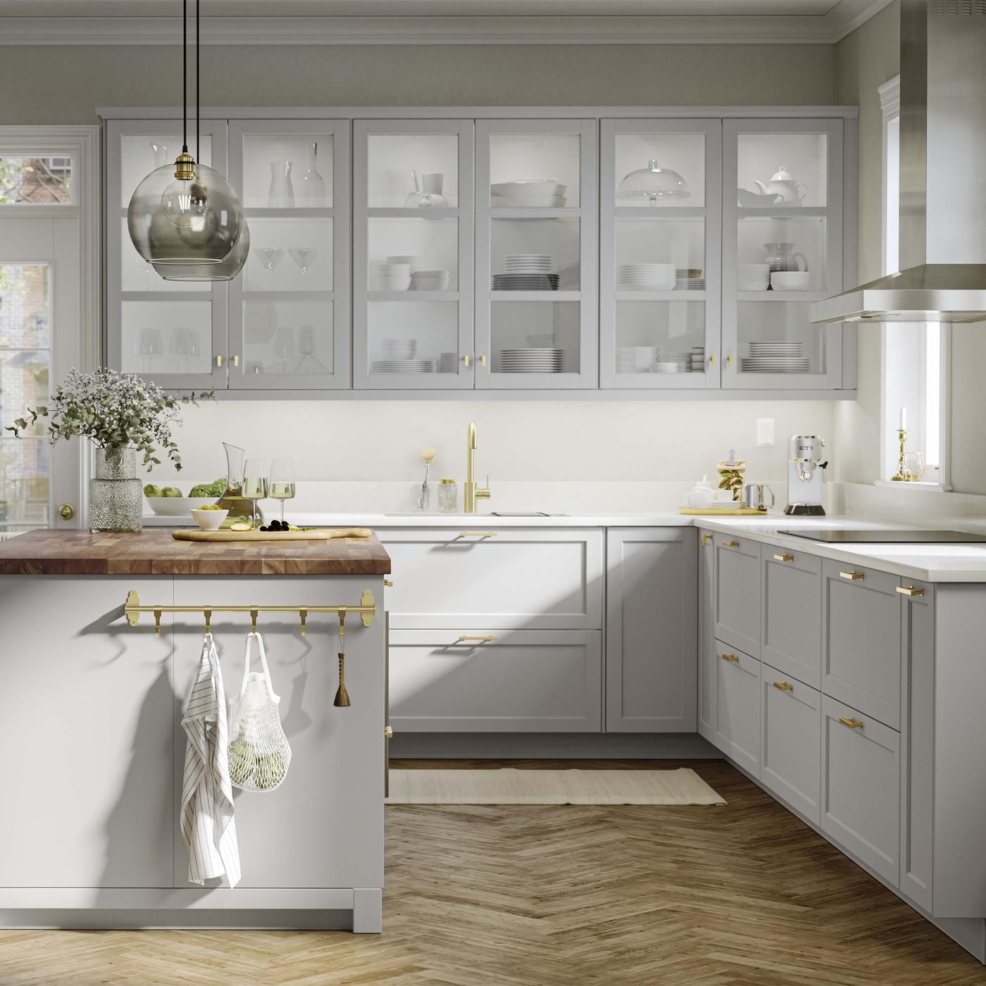 Bright modern kitchen with light gray cabinets, glass uppers, gold hardware and a wood‑topped island with hanging towels.
