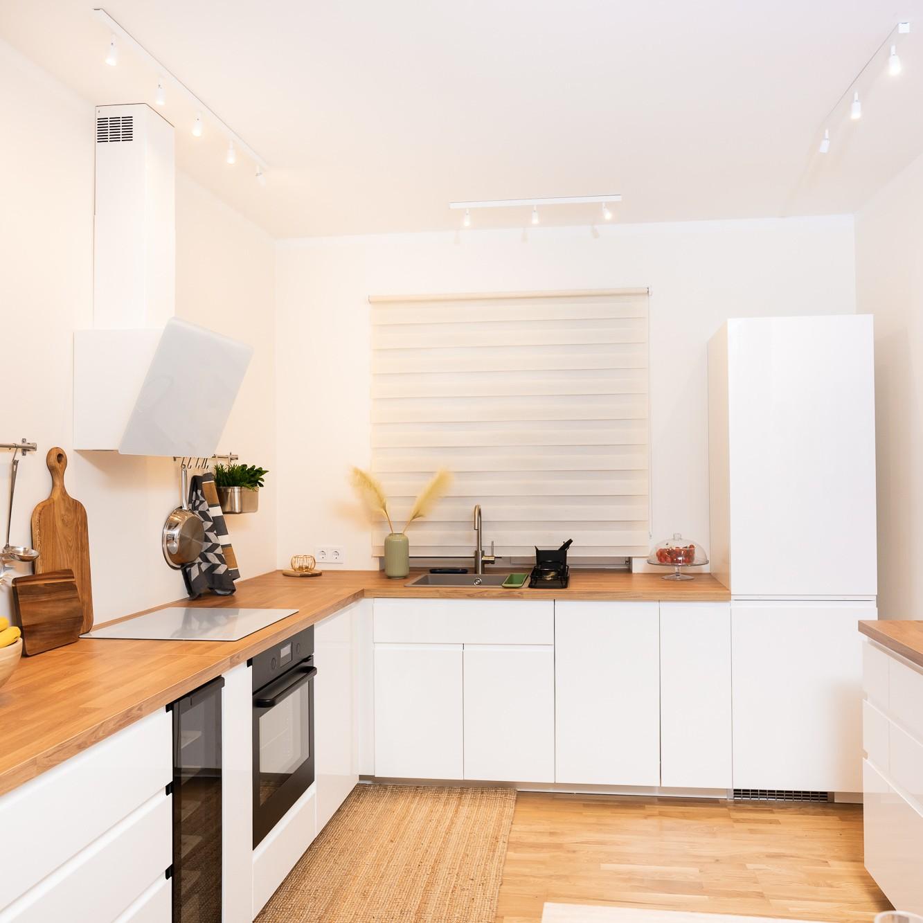Bright, modern L-shaped kitchen with white cabinet fronts and wooden worktops; kitchen utensils and plants placed on the counter, creating a tidy and spacious look.