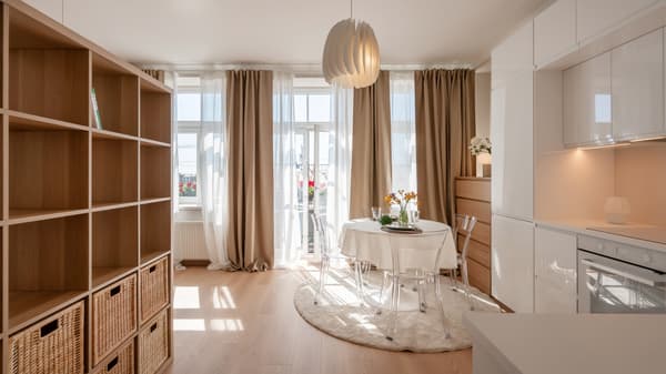 Bright modern studio flat with light wood flooring, white furniture, a small dining table, and large windows with beige curtains.