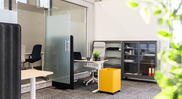 Bright office workspace with ergonomic chair, yellow storage bin, and glass meeting room.