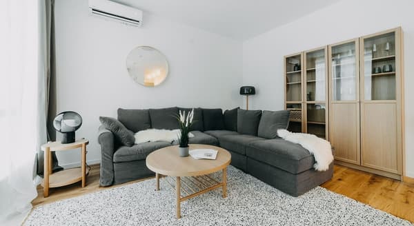 Bright Scandinavian-style living room with a large grey sectional sofa, light wood coffee table, glass-door cabinet, round wall mirror, indoor plant decor, and minimalist lighting on wood flooring with a white speckled rug.