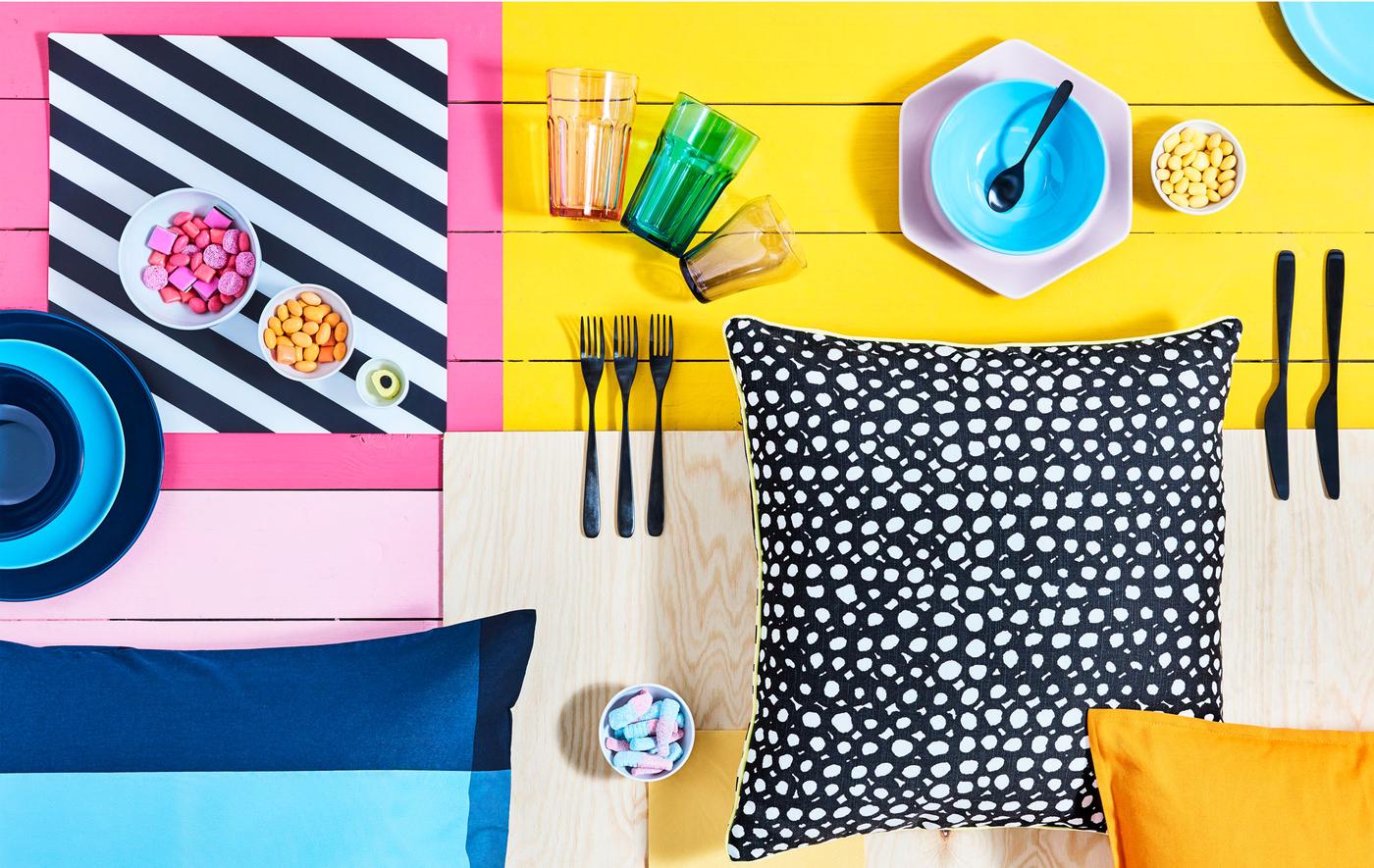 Brightly block-colored surface with various tableware, bowls of candy and patterned cushions laid out on it.