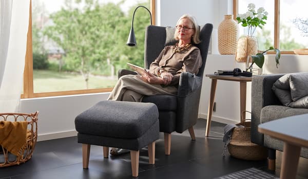 Britt Monti assise dans un fauteuil OMTÄNKSAM dans le coin lumineux d’une pièce avec de grandes fenêtres donnant sur un jardin.
