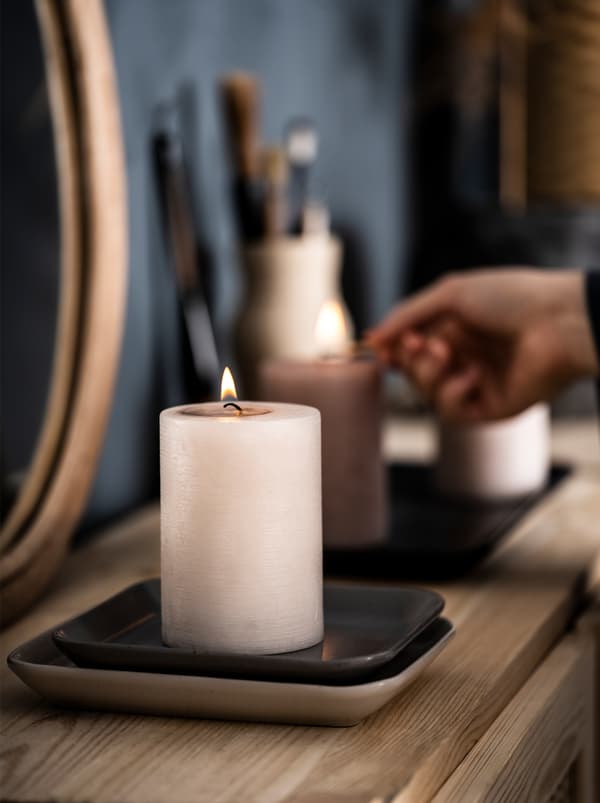 Buffet in legno in una camera da letto, con specchio e oggetti decorativi. Una mano accende due ceri profumati FÖRSÖKA.