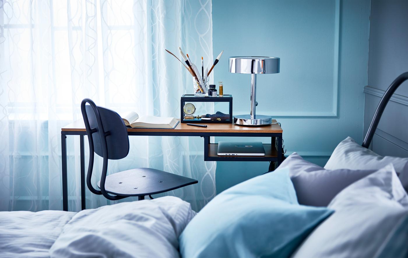 Bureau, sur roulettes, en bois massif et en métal, et chaise pivotante installés dans une petite chambre, entre le mur et le lit