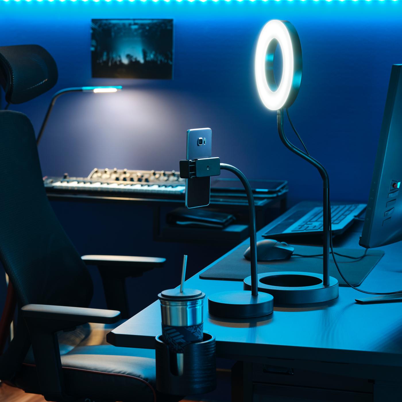 ブルーに照らされたゲーム部屋。ゲーム用デスクの上にLÅNESPELARE/ローネスペラレ リングライト 携帯電話ホルダー付きが置いてある。その手前にMATCHSPEL/マッチスペル ゲーム用チェア。
