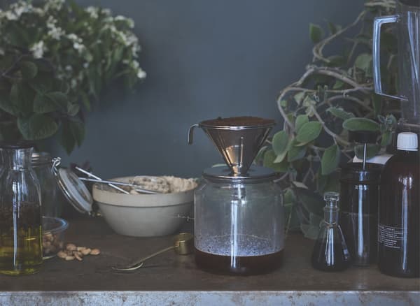 Café en train de s’écouler dans une carafe en verre, posée sur une table avec d’autres bocaux et ingrédients.