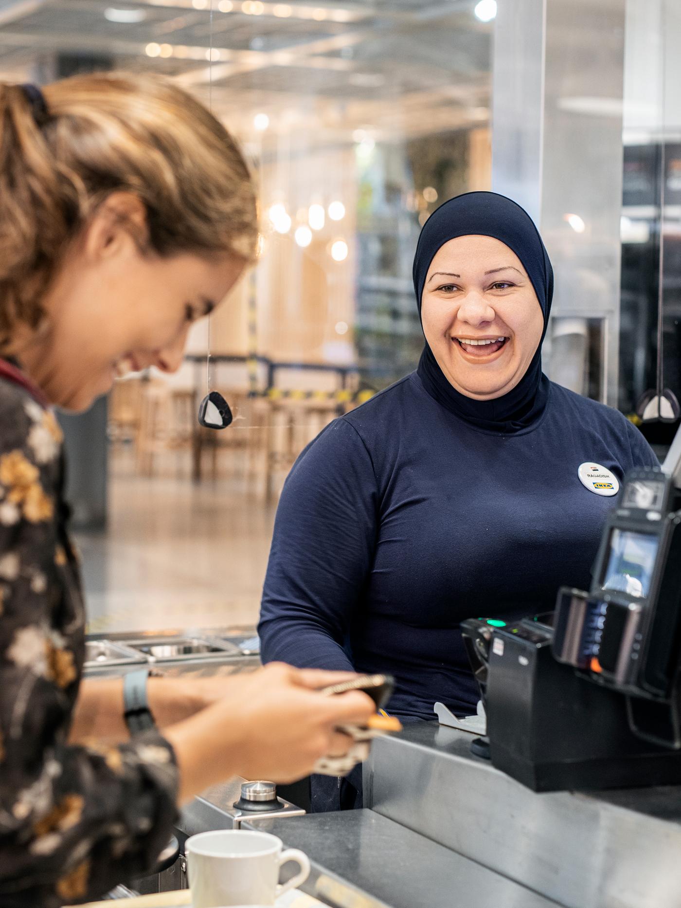 Caissière IKEA avec un hijab partageant un bon moment avec une cliente à la caisse.