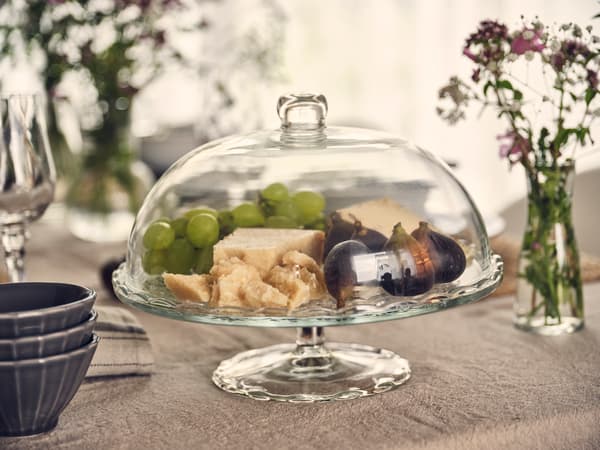 Campana di vetro con formaggio, uva e fichi su una tavola imbandita, impreziosita da fiori e stoviglie.