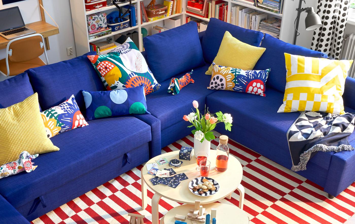 Canapé d’angle FRIHETEN bleu, cousins colorés, deux tables basses rondes et tapis rayé rouge et blanc