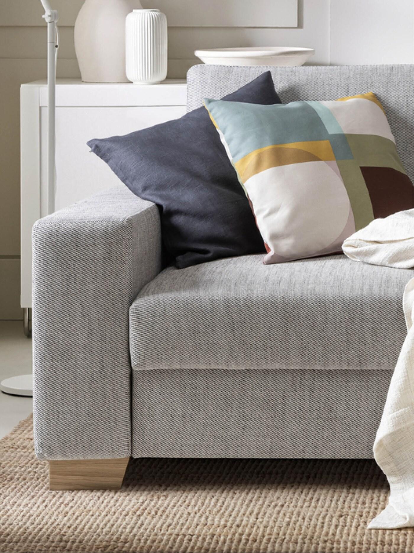 Canapé gris avec coussin bleu foncé et coussin à motif géométrique coloré, couverture blanche sur l'accoudoir, sur tapis beige avec armoire et décoration blanches en arrière-plan.