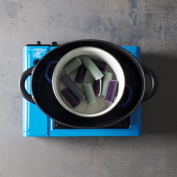 Candle remnants in various colors are heated in a small pot au-bain-marie inside a larger pan, as if they were being melted down for an IKEA-style DIY candle project.
