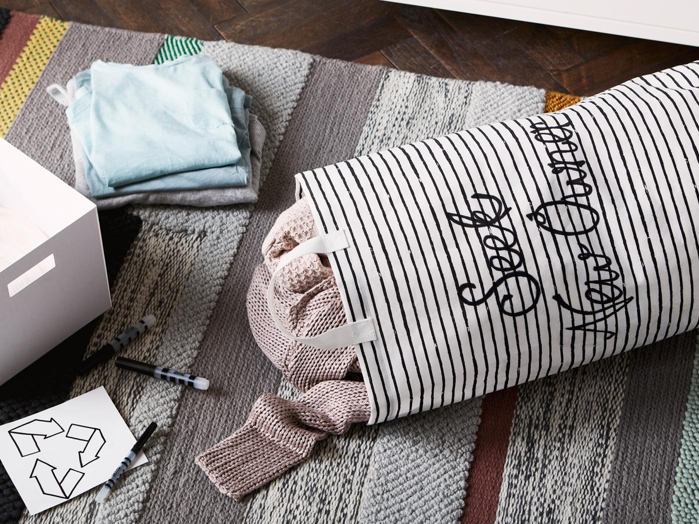 Canvas tote bag embroidered with the words ‘Seek new owner’ and clothes inside lying on a rug next to a box, pens and recycling label.