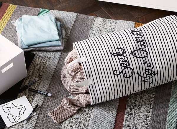 Canvas tote bag with words ‘seek new owner’ and clothes inside lying on a rug next to a box, pens and recycling label.