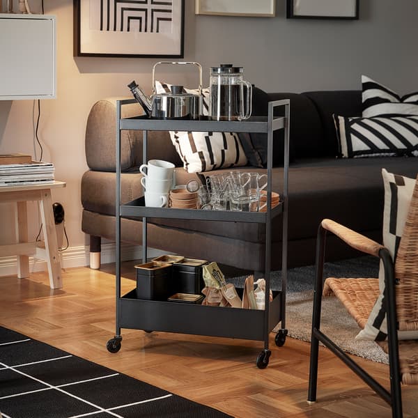 Carrello NISSAFORS nero con un bollitore, delle tazze e delle bustine di tè, posizionato accanto a un divano e a una poltrona - IKEA