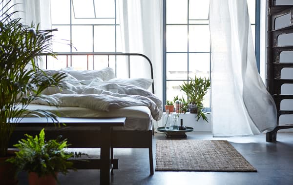 Chambre à coucher avec lit défait, fenêtres ouvertes, tapis sur le sol et un grand nombre de plantes vertes