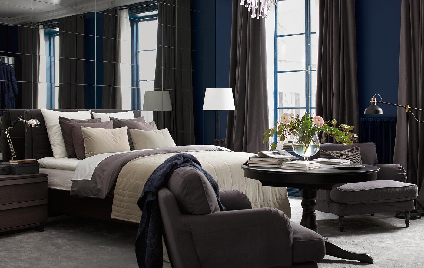 Chambre avec mur de miroirs, lit garni de nombreux textiles, fauteuil, lampadaires et fleurs coupées.