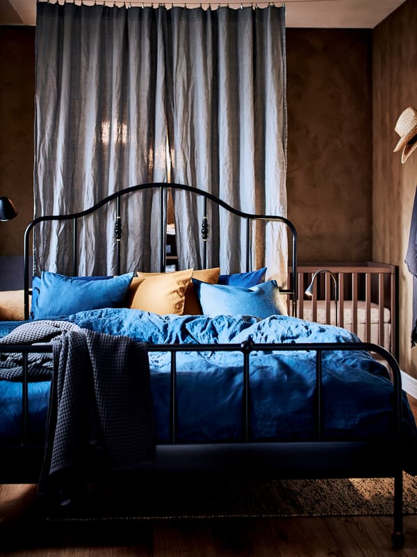 Chambre avec un lit SAGSTUA noir; dans un coin, derrière un rideau servant de séparateur, se trouve un lit bébé en bois.