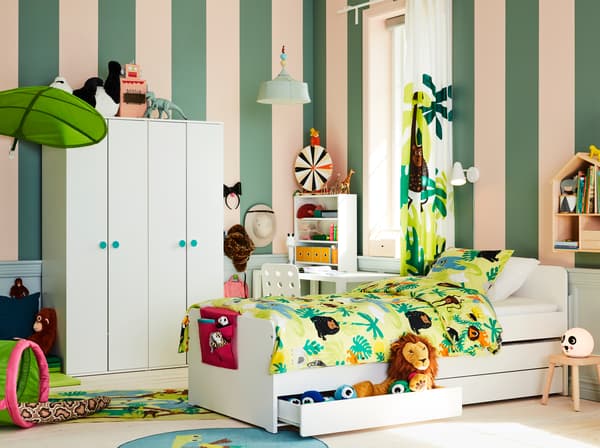Chambre d’enfant avec déco façon jungle, cadre de lit blanc, peluches, textiles à motifs animaux et ciel de lit en forme de feuille.