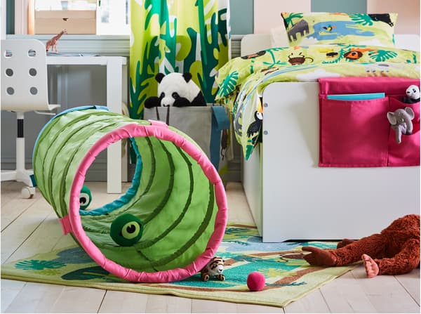 Chambre d’enfant avec tunnel de jeu vert installé sur un tapis à motifs jungle. Il y a des peluches dans le tunnel et autour de celui-ci.