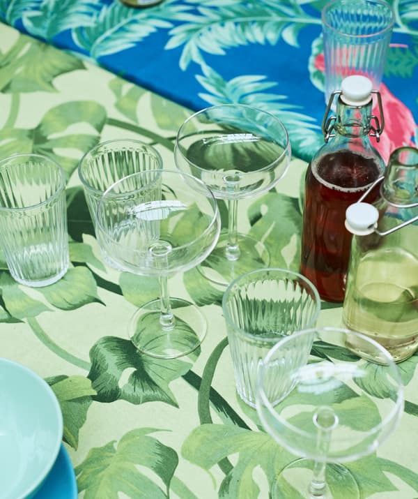 Champagne glasses and drinking glasses, with glass jars filled with drink on a table covered with green leaf-print fabric.