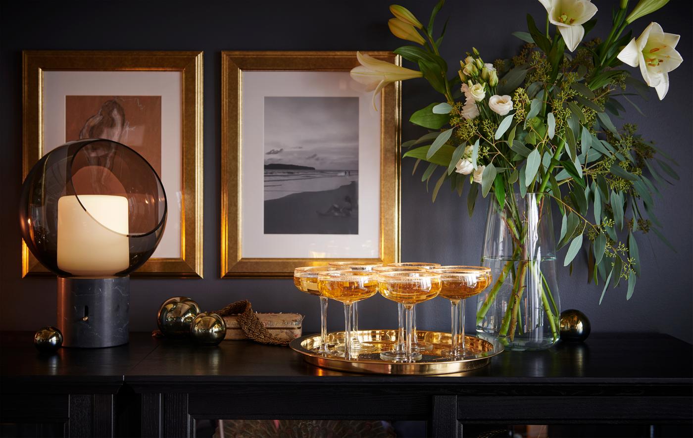 Champagne glasses in a golden tray on a black table between an EVEDAL lamp and a vase of flowers.