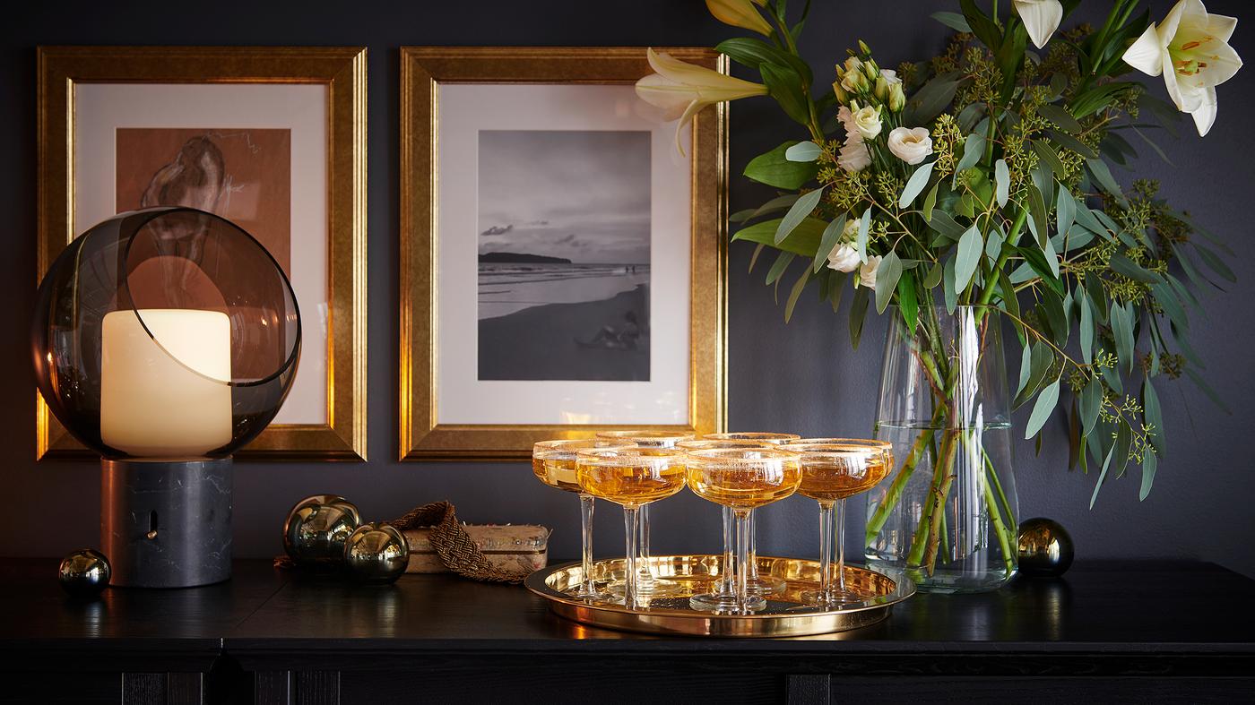 Champagne glasses on a tray, in front of a flower arrangement in a glass vase and two gold-framed pictures on the far wall.