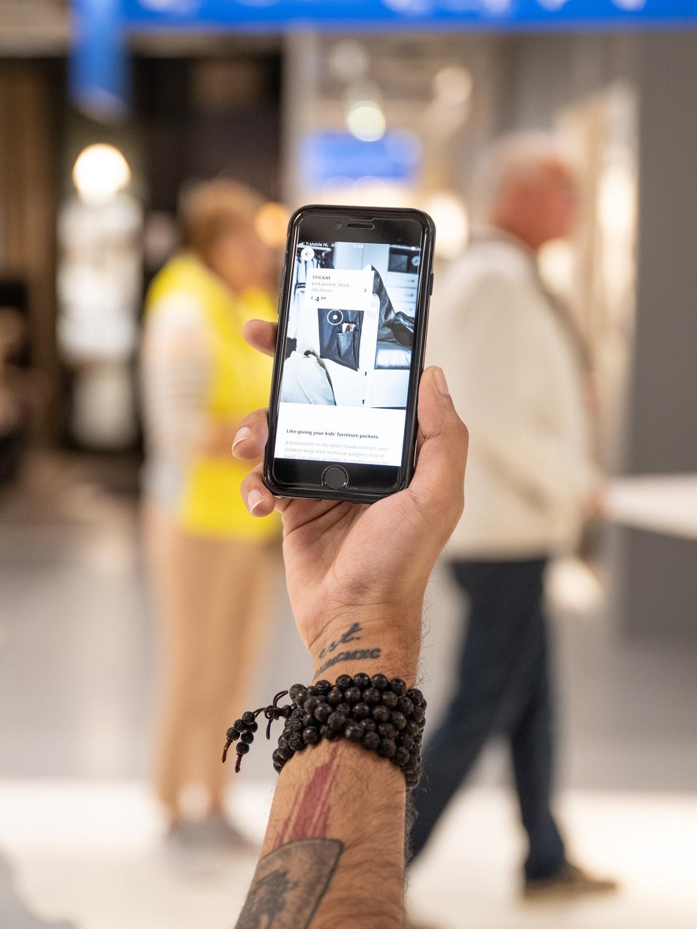 Человек стоит в магазине и держит смартфон с открытым приложением IKEA; на заднем плане видны другие посетители.