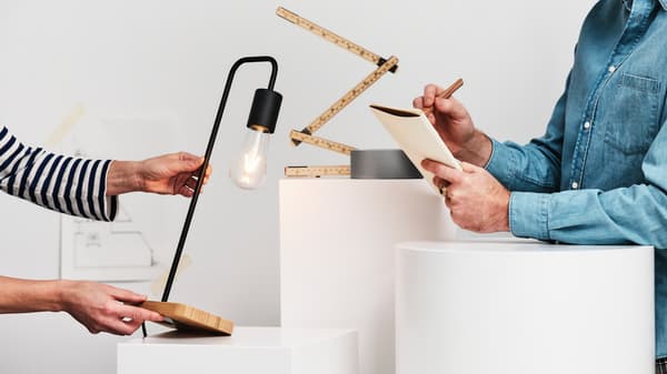 Человек в синей джинсовой рубашке пишет в блокноте, а другой держит черную/бамбуковую настольную лампу TVÄRHAND.