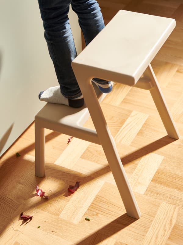 Child standing on the lower step of a MÄSTERBY step stool, placed next to a kitchen. Red-onion scraps on the floor.