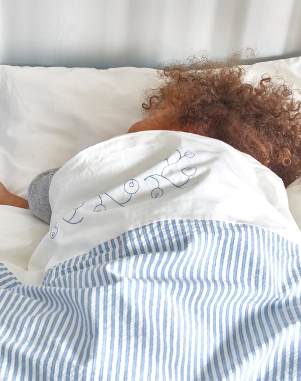 Child with brown curly hair lying asleep in a bed with GULSPARV blue and white striped bed linen duvet and white pillow.