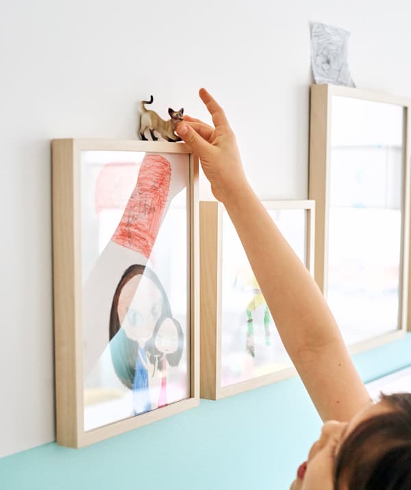 Children’s drawings in light wood frames, wall-mounted in a line. A girl balances a cat figurine on top of the first frame.