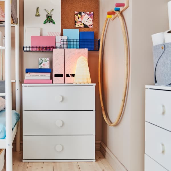 Children's room with two white chests of drawers that have books, a white wall lamp and pink magazine files standing on top.