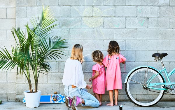 Chloé and her daughters drawing in chalk on a grey brick wall, with a pot plant and blue bike.