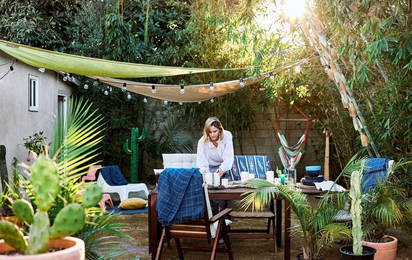 Chloé en train de dresser la table dans le jardin, avec des meubles en bois, des plantes, des arbres et des auvents avec guirlandes lumineuses.