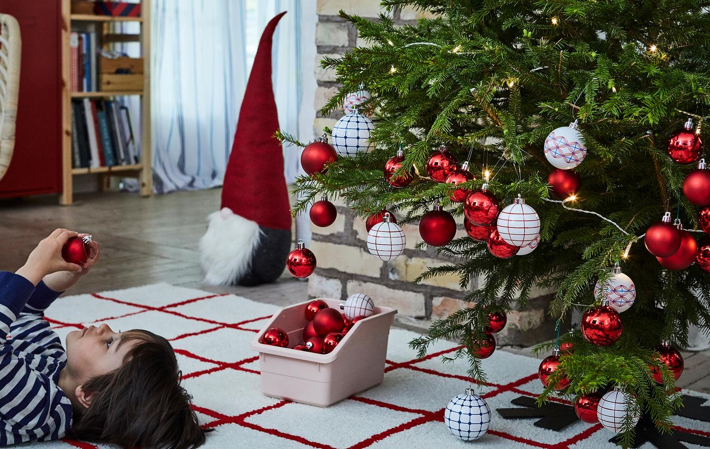 Christmas tree heavy with decorations with a young boy lying face up on rug next to it, inspecting an ornament.