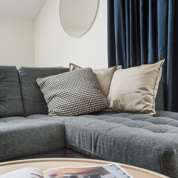 Close-up of a cozy corner of a dark gray sectional sofa with two large throw pillows—one beige and one patterned in black and white. Behind the sofa, deep navy-blue curtains cover a window, and a round wall mirror hangs on the white wall. A light wood coffee table with a partially visible open magazine is in the foreground. The scene conveys a modern, soft, and inviting atmosphere.
