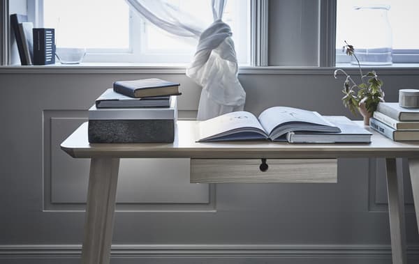 Close-up of a desk with open books on it, in front of a window with a knotted curtain.