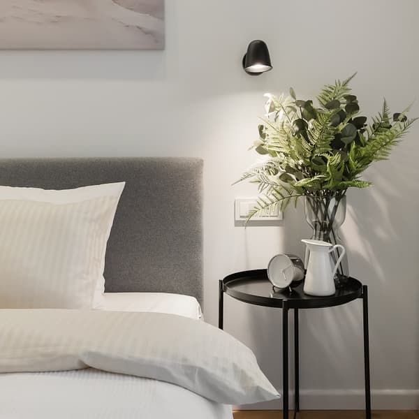 Close-up of a modern bedside setup with a grey upholstered bed and cream-striped bedding. A black round side table holds a white ceramic jug, a classic alarm clock, and a clear vase with vibrant green foliage. A minimalist black wall sconce casts soft light on the arrangement.