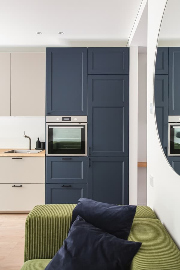 Close-up of a modern kitchen corner featuring deep navy blue cabinetry with integrated oven and tall storage units. Light beige lower cabinets with a wood countertop and matte black hardware contrast elegantly with the darker elements. Part of a green corduroy sofa with navy throw pillows is visible in the foreground, alongside the edge of a round wall mirror, reflecting the cohesive and stylish design of the living space.