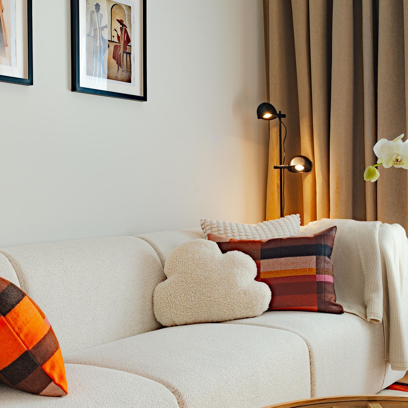 Close-up of a natural white sofa with textured cushions in terracotta tones, framed artwork, soft curtains, and a floor lamp creating a warm, calm living area.