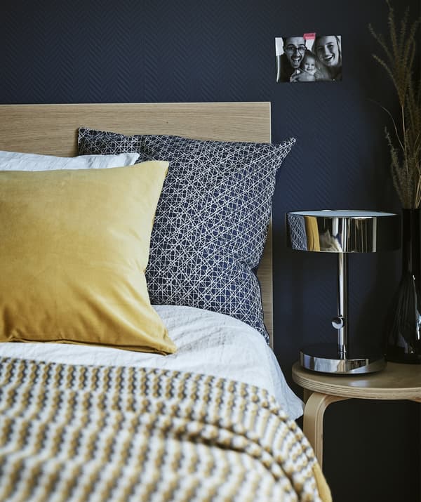 Close up of the corner of a bed, bedside table and lamp against a dark blue wall.