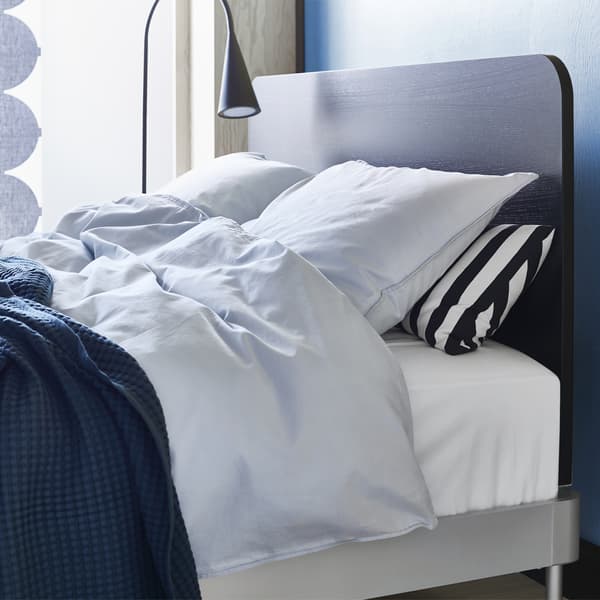 Close-up of the corner of a double bed with a black headboard, floor lamp and light bedding.