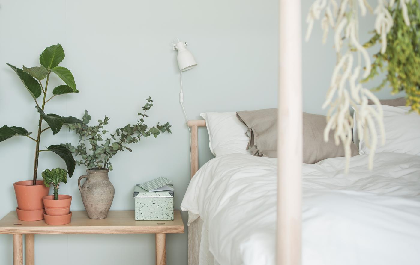 Close-up van een GJÖRA berken bed met wit textiel en een essen nachtkastje met sierpotten van aardewerk en groene planten