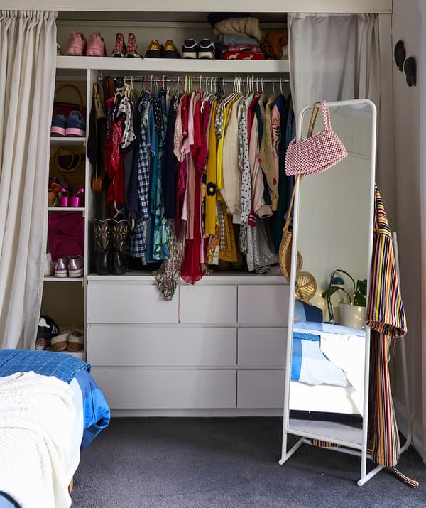 Clothes hanging on a rail and shoes on shelves above two chest of drawers, with curtains either side and a free-standing mirror.