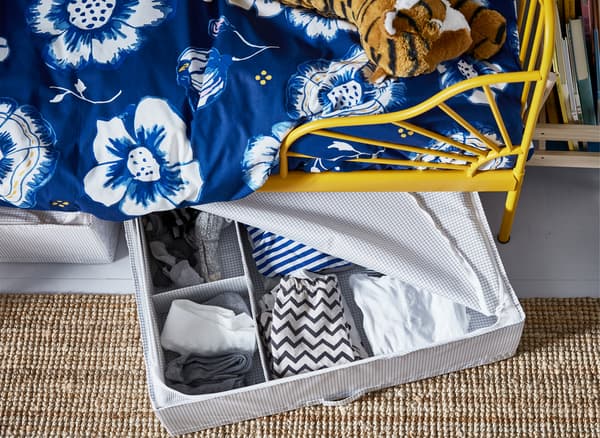 Clothes in an open storage box under a child’s bed with yellow frame and blue floral bedding.
