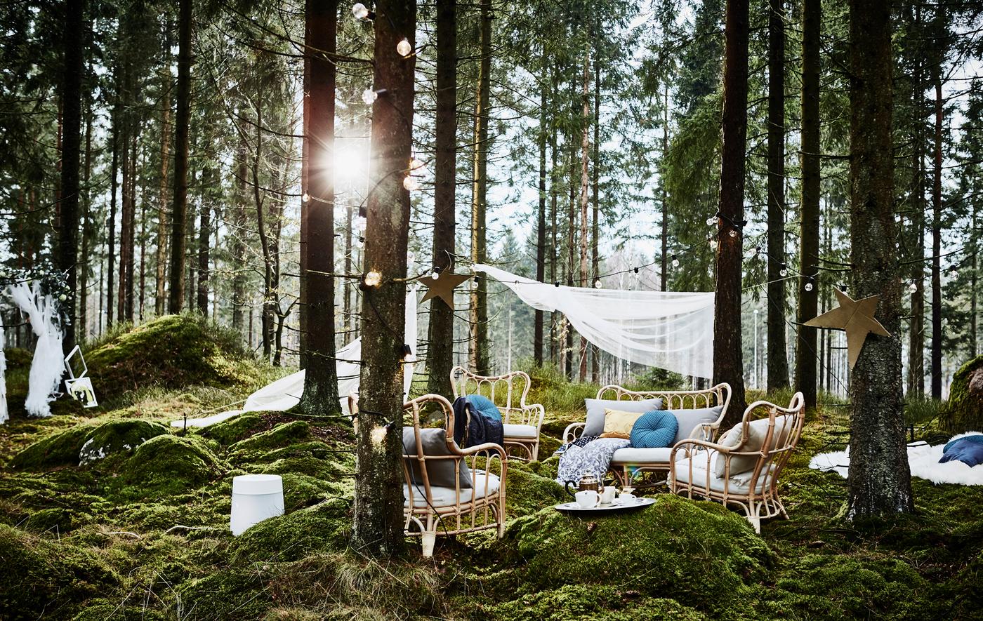 Coin détente lors d’une fête de noces organisée en forêt, avec fauteuils en rotin, nombreux coussins et guirlandes lumineuses, à côté d’un photomaton maison